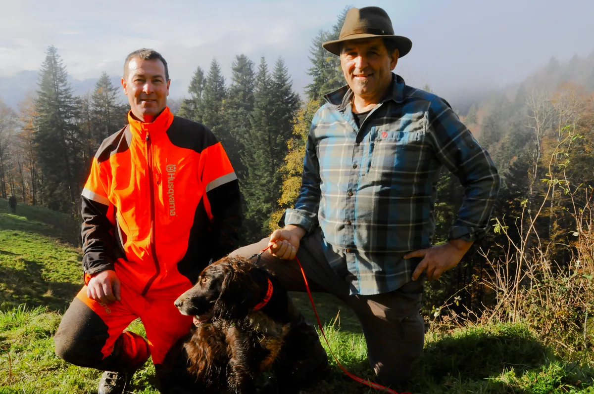 Man sieht zwei Jäger mit einem Jagdhund.